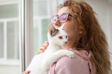 Attractive girl in pink clothes and eyeglasses with withe cat in her arms standing near the window at home. Pet owner concept