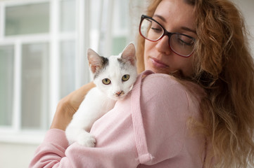 Attractive girl in pink clothes and eyeglasses with withe cat in her arms standing near the window at home. Pet owner concept