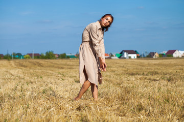 Naklejka na ściany i meble Young beautiful brunette model in a beige dress posing against the background of a mowed wheat field