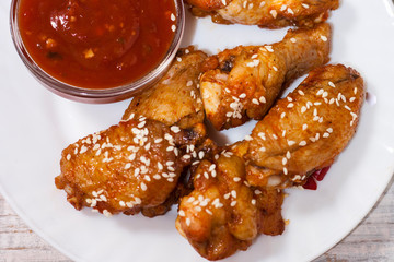 Fried juicy chicken wings marinated with honey, soy sauce, spices, sprinkled with sesame seeds on a white plate with sauce. Asian recipe, top view, close up