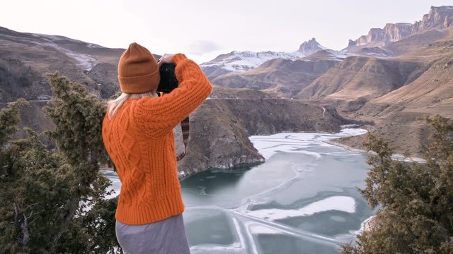 Back view Girl photographer traveler in an orange sweater and hat takes pictures on his dslr camera landscape on the outdor against the backdrop of epic mountains. Travel Photographers Video Concept