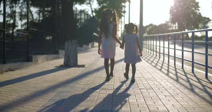 Back view two pretty sisters seven and four years old walk holding hands along a city street of paving slabs with a metal fence along the road. Sunny summer evening. Slow Motion 4k 50 fps