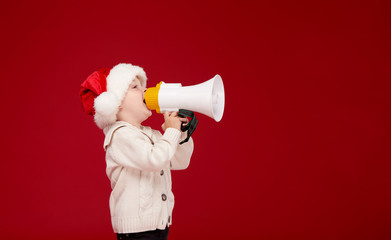 Happy child boy in santa hat. Kid shouting by megaphone at Christmas night. Happy xmas and New Year, winter holiday concept with copy space for text on red background