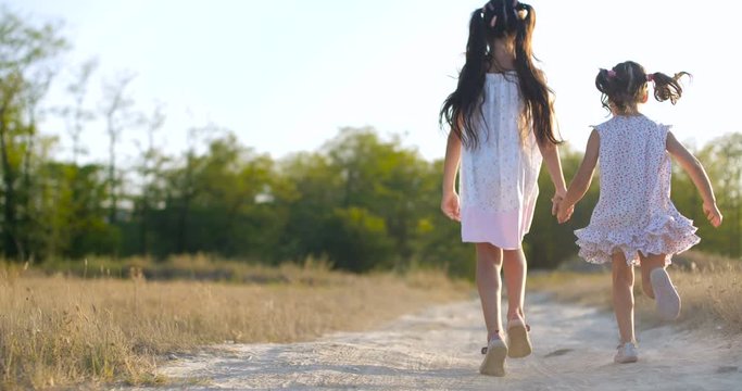 Back view  close up two beautiful little girls of four and seven years old in summer dresses running dusty country  trail in a meadow  on warm summer evening. Slow motion 50 fps 4k