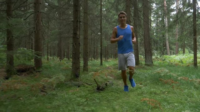 SLOW MOTION: Sweaty athletic man running through the idyllic green forest as he prepares for a trail running competition. Cheerful Caucasian man jogging down an empty path crossing the scenic woods.