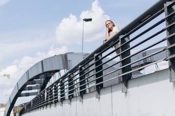 Outdoors urban portrait of young hipster blondie woman in glasses with long  hair smiling and sitting on bridge. Architecture, urban style, megalopolis concept