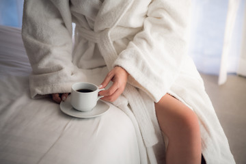 Close-up of legs of woman in white terry bathrobe on bed with cup of coffee in the morning. Ideal...