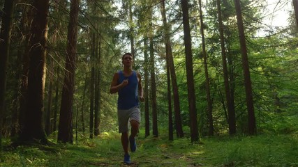 LOW ANGLE, CLOSE UP, SLOW MOTION: Energetic athletic man goes trail running through the scenic forest on a sunny spring afternoon. Carefree Caucasian male athlete jogging through the sunlit woods.
