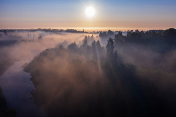 The rays of the morning sun make their way through the thick fog over the forest, field and river