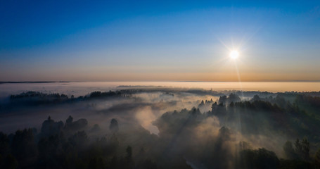 The rays of the morning sun make their way through the thick fog over the forest, field and river