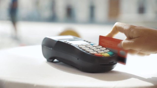 Girl paying in the cafe by credit card. Young woman close-up swiping with credit card on pos payment terminal on table in sunlight.
