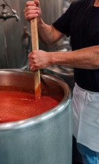 person stirring red sauce in an industrial size pot, man in an apron stirring a vat of tomato sauce with a wooden paddle in a commercial kitchen