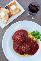 spaghetti with red sauce and meatballs with a sprig of parsley and grated parmesan in a white bowl, fresh made Italian pasta on a stone table with a glass of red wine and buttered toast