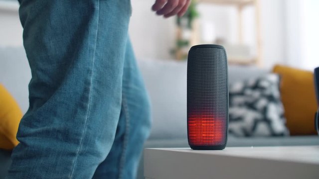 Man listens to music from a portable speaker that glows in different colors in the living room. 4k slow motion footage.