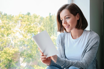 Fototapeta premium Beautiful young woman reading book near window