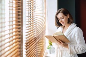 Morning of beautiful young woman reading book near window