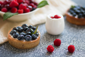 Tartalets with blueberry berries, plate with fresh raspberry and dairy with sour cream on the grey surface. 