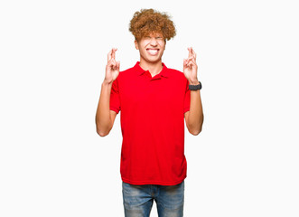 Young handsome man with afro hair wearing red t-shirt smiling crossing fingers with hope and eyes closed. Luck and superstitious concept.