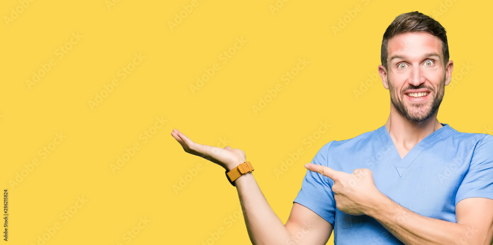 Wall mural Handsome doctor man wearing medical uniform over isolated background amazed and smiling to the camera while presenting with hand and pointing with finger.