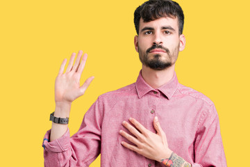Young handsome man wearing pink shirt over isolated background Swearing with hand on chest and open...