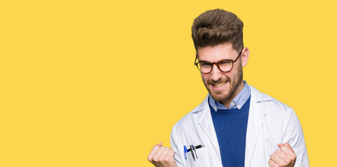 Young handsome scientist man wearing glasses very happy and excited doing winner gesture with arms raised, smiling and screaming for success. Celebration concept. - obrazy, fototapety, plakaty