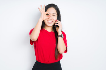 Young brunette woman talking on smartphone over isolated background with happy face smiling doing ok sign with hand on eye looking through fingers