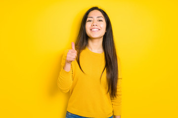 Beautiful brunette woman over yellow isolated background doing happy thumbs up gesture with hand. Approving expression looking at the camera with showing success.