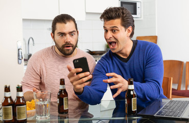 Friendly male meeting over beer at home, men looking at phone and discussing