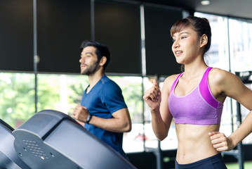 Fototapeta na wymiar Young fit beautiful Asian woman and handsome Caucasian man running on treadmill or running machine in modern fitness gym. Seen from side view while they focusing on running. Workout in Gym and fitness