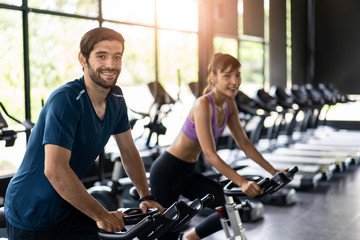 Fototapeta na wymiar Young fit handsome Caucasian man and beautiful Asian woman cycling on bike machine in modern fitness gym. Seen from side view while they focusing on exercising. Workout in Gym and fitness for health.
