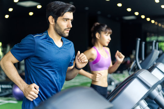 Young fit handsome Caucasian man and beautiful Asian woman running on treadmill or running machine in modern fitness gym. Seen from side view while they focusing on running. Workout in Gym and fitness