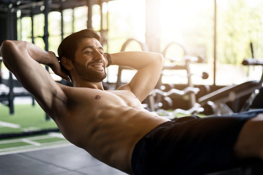Young handsome smiling Caucasian man with beard and shirtless exercising by doing a sit-up on bench in gym or fitness club. He has a good body shape with six pack. Sport recreation and fitness concept