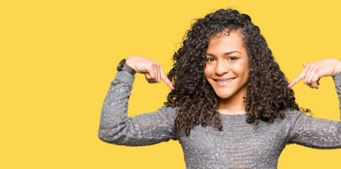 Young beautiful woman with curly hair wearing grey sweater looking confident with smile on face,...