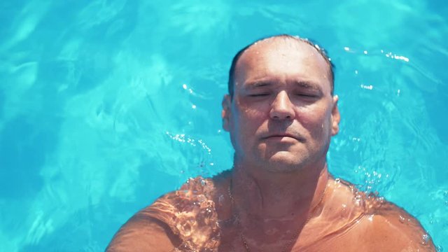 adult relaxed man chilling in the pool's blue tranquill water. Handsome man lying on the water in the pool close up in Slow motion