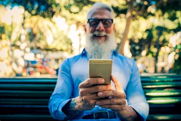 Trendy senior man using smartphone app in the park. Mature fashion male having fun with new trends technology . Tech and joyful elderly lifestyle concept - Focus on his hands