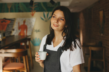 Fototapeta na wymiar Portrait of smiling charming brunette girl with braces on teeth in casual clothes with cup of coffee in cafe
