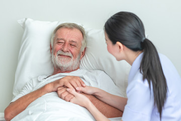 Obraz premium Senior man in bed with nurse in retirement home. Caucasian male with asian woman. Happy smile.