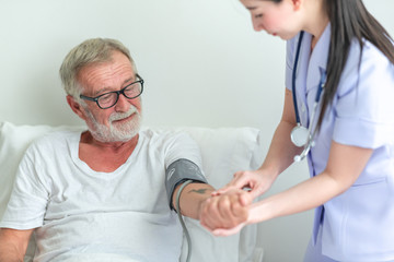 Obraz premium Senior man in bed with nurse in retirement home. Caucasian male with asian woman. Using blood pressure monitor.