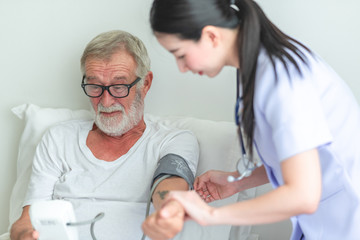 Senior man in bed with nurse in retirement home. Caucasian male with asian woman. Checking result of blood pressure.