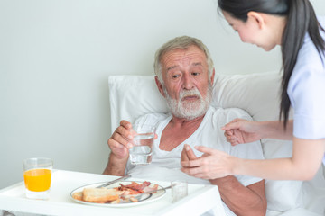 Senior man in bed having medication with nurse. Caucasian man with asian woman.  Talking.