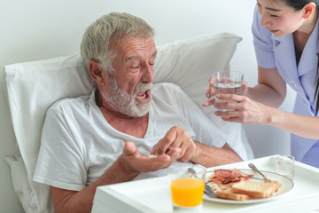 Senior man in bed having medication with nurse. Caucasian man with asian woman.  Scare of medical pill.