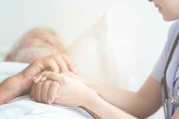 Senior man in bed with nurse in retirement home. Caucasian male with asian woman. Holding hands...