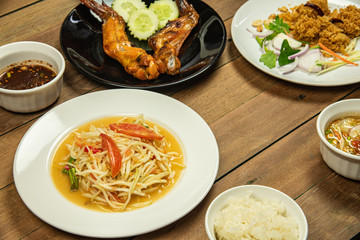 Most popular Thai northeast food with papaya salad, grilled chicken wings, fluffy deep fried catfish and spicy mango sauce on wooden table background.