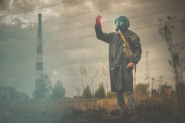Man in gas mask is looking a radioactive liquid in vial in his hand on a smoking chimney...