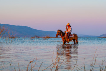 a man cowboys is riding horse back in a water with mountain during sunset