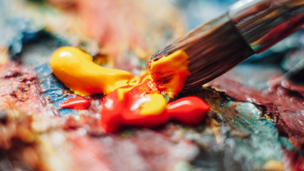 Creative artist using paintbrush to mix yellow and red oil paint on colorful palette.