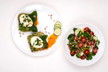 Sandwiches with poached eggs and salad with avocado, cucumbers, cherry tomatoes, spinach leaves, parsley and seeds. Breakfast food on a white background. Top view.