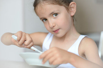 beautiful little caucasian girl in white tank top eating breakfast holding plate and spoon sitting in bright kitchen