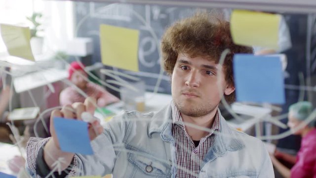 Chest-up portrait shot of young Caucasian male creative agency specialist with curly hair, in denim jacket, drawing diagram on glass panel and planning tasks for project, then looking at camera