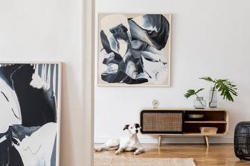 Design scandi home interior of living room with wooden commode, gray sofa, black rattan pouf, plant and elegant accessories. Stylish home decor. Mock up abstract paintings.  Dog is lying on the carpet